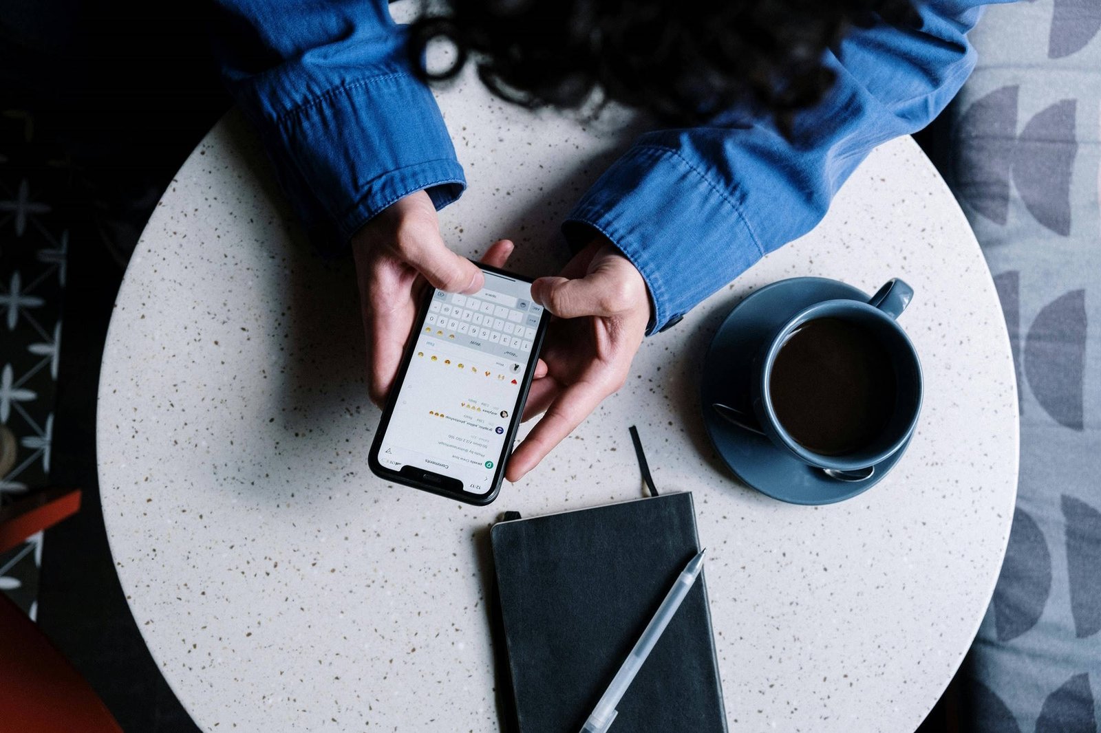 Overhead view of a person in a blue denim shirt sitting at a white speckled round table, holding a smartphone displaying a messaging app with emoji keyboard. A blue coffee cup and black notebook with pen are placed on the table beside them.