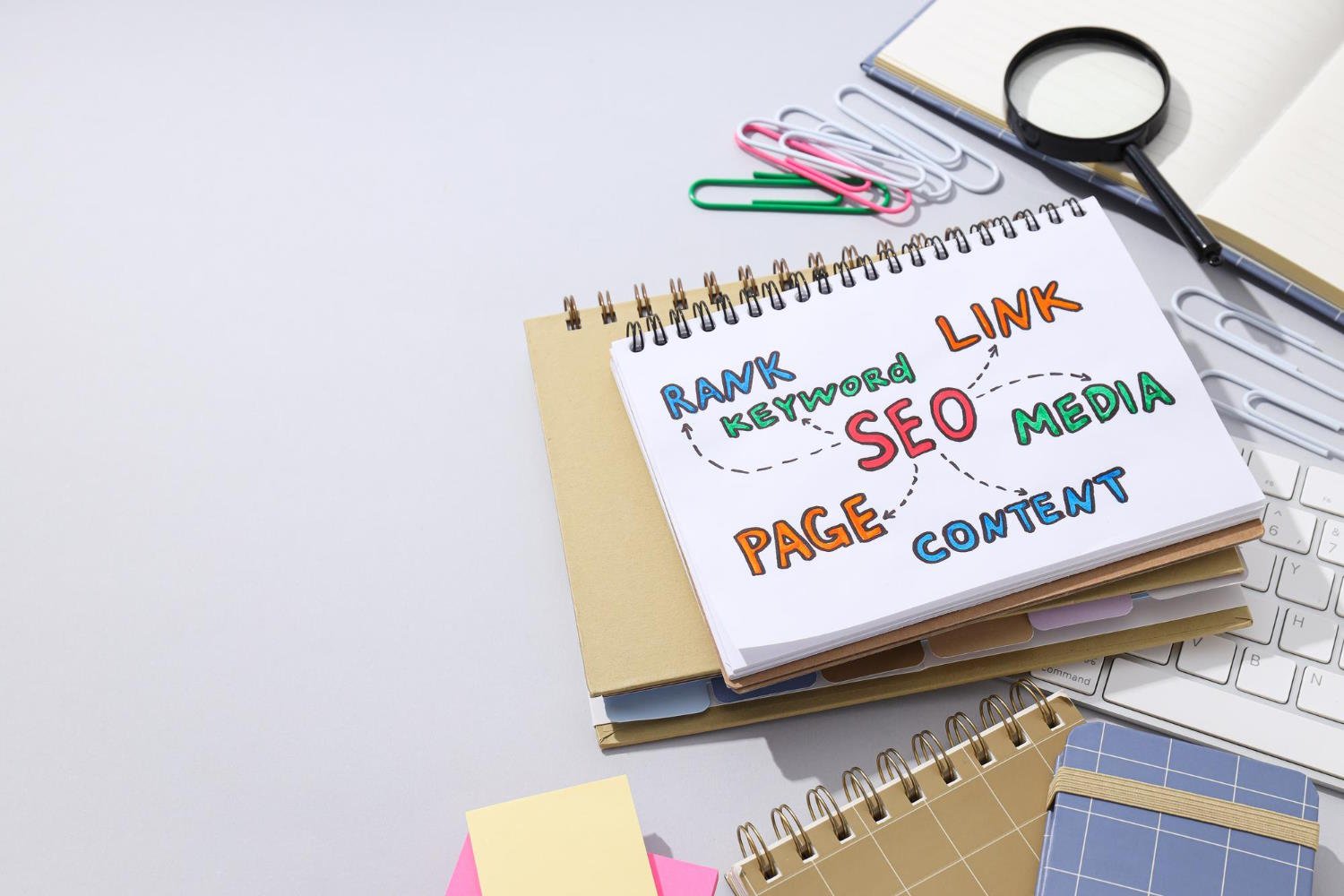 SEO strategy planning workspace showing handwritten notes with keywords like RANK, KEYWORD, SEO, LINK, social MEDIA, PAGE, and CONTENT connected by arrows on a spiral notebook, surrounded by office supplies including paper clips, magnifying glass, and keyboard